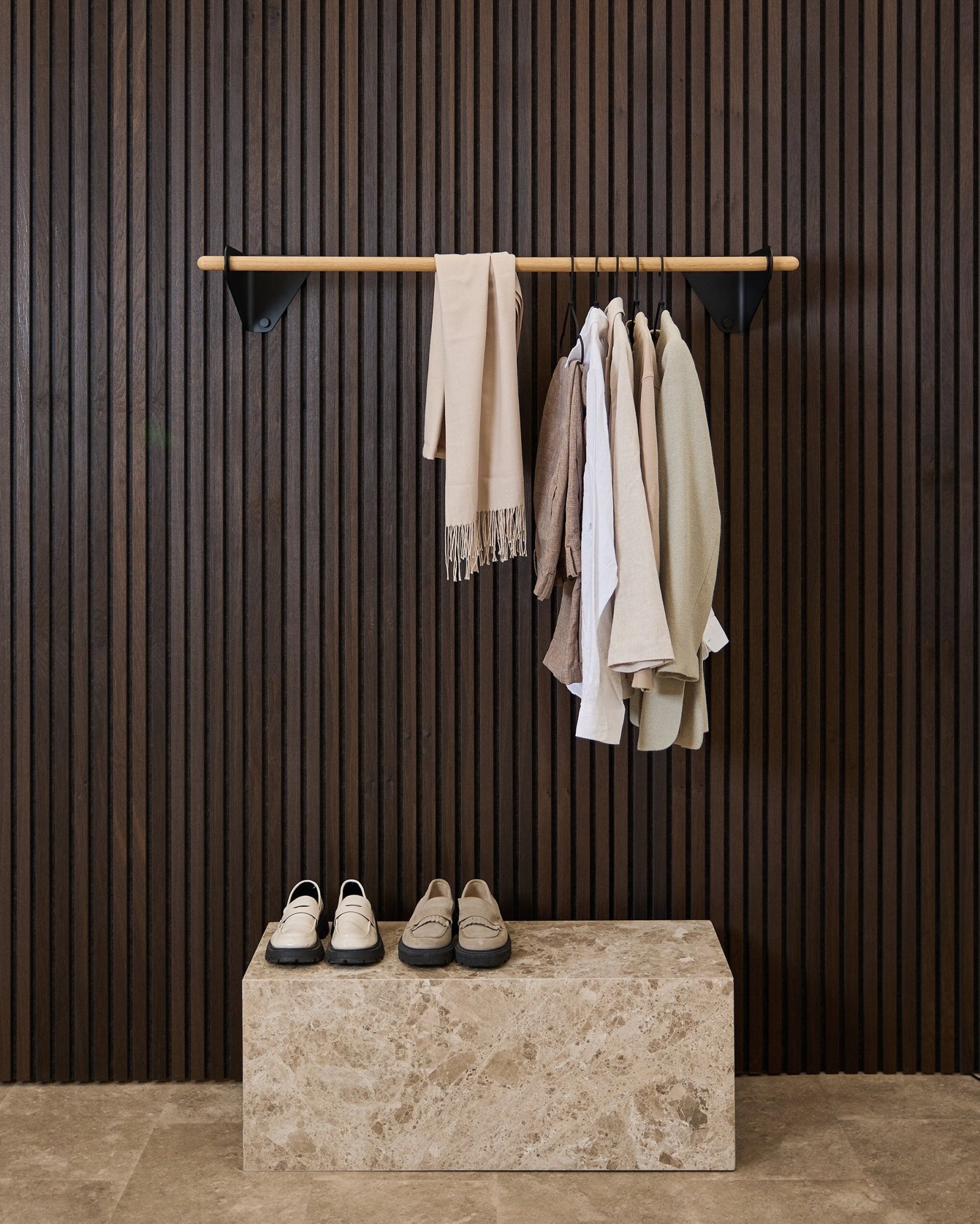 Neutral-toned clothes and a scarf hang on a black 115.9 cm Linn | Coat Rail by WoodUpp above a marble bench, against a dark Akupanel acoustic slatwall panel.